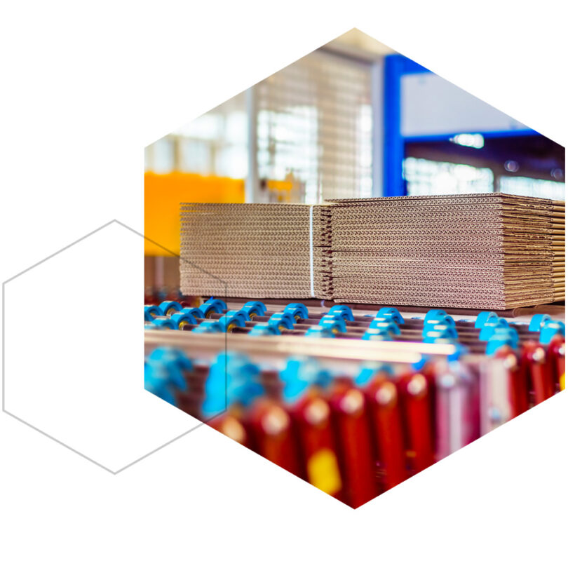 Stacked corrugated cardboard sheets on a conveyor system inside a packaging or manufacturing facility, shown within a hexagonal crop.