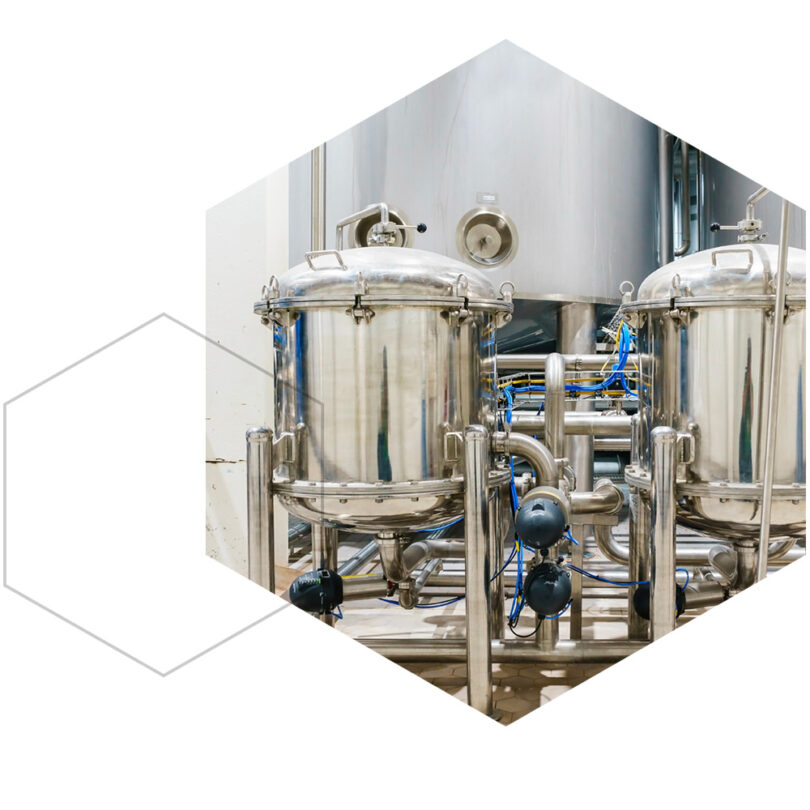 Two large stainless steel tanks with attached pipes and valves in a clean industrial processing facility, framed in a hexagonal crop.