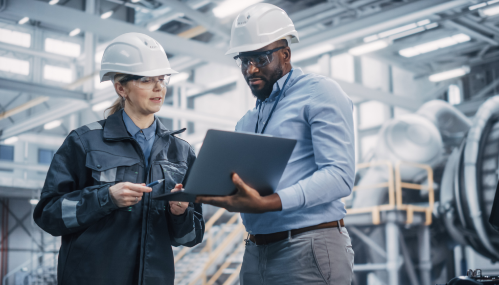 Two industrial professionals in hard hats reviewing data on a laptop in a modern manufacturing facility.
