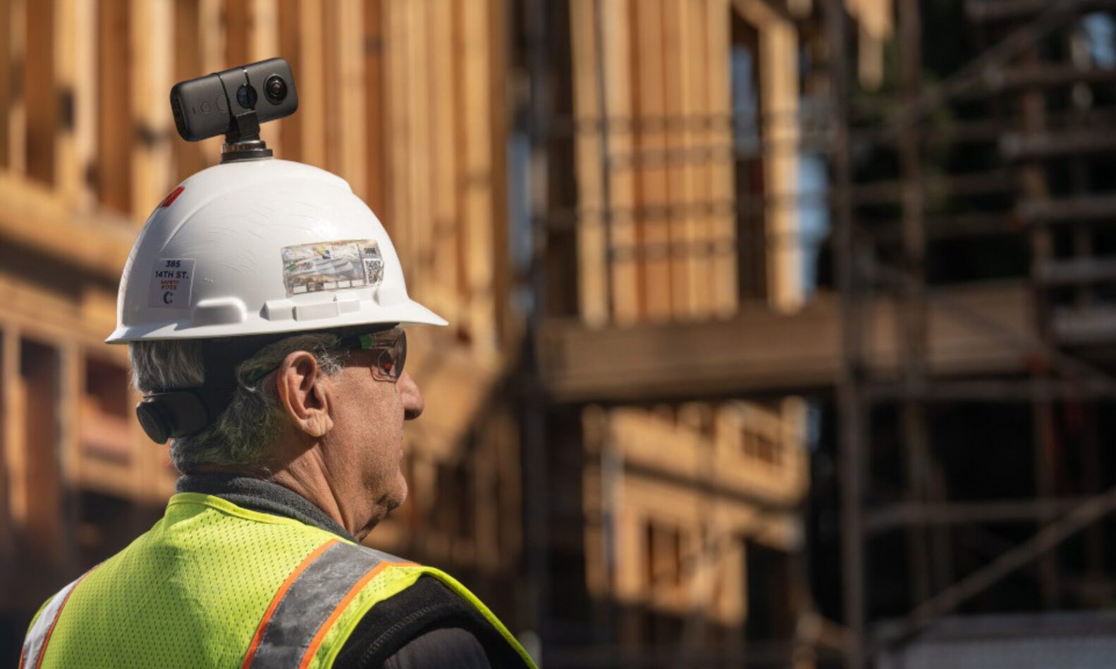 360 camera mounted on a helmet | prevu3d Construction worker in a neon safety vest and white hard hat with an action camera mounted on top, at a wooden-frame construction site.