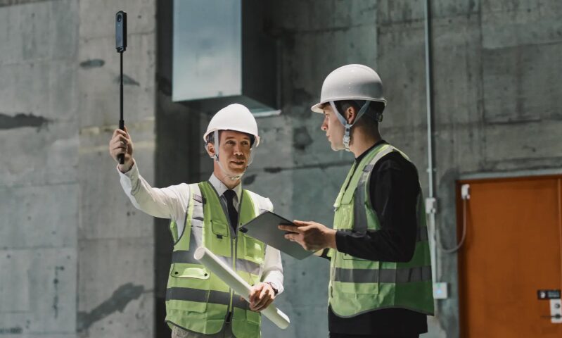 360 capture | prevu3d Two construction workers in hard hats and hi-vis vests discuss plans; one holds a blueprint and a selfie stick, the other a clipboard.