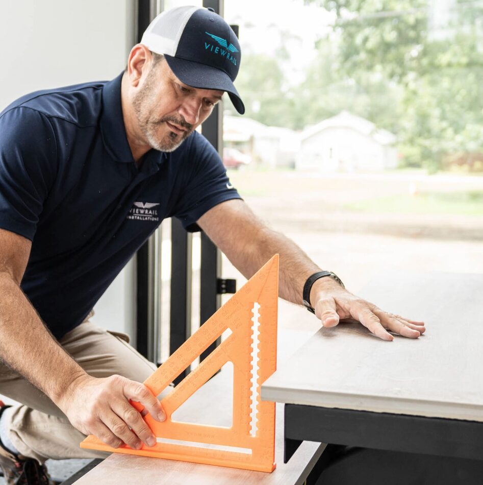 viewrail thumbnail | prevu3d Carpenter in a navy polo and cap measures a wooden board with an orange speed square.