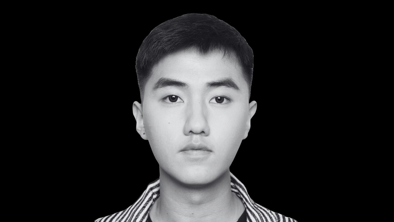 Javier Zhang | Prevu3d Portrait of a Young Person Facing the Camera Head and shoulders Shot in Black and White Against a Black Background Wearing a Striped Collar Shirt Prevu3d