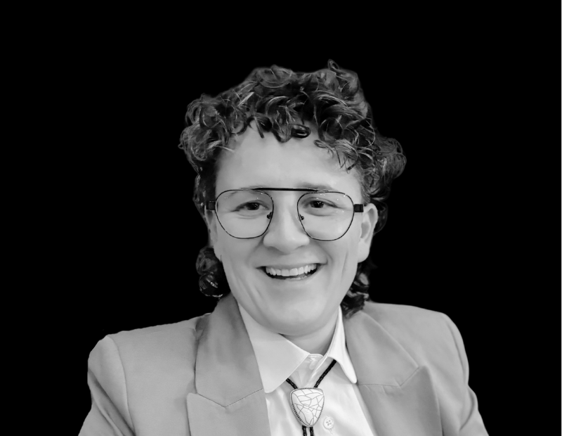 Elle Magni | Prevu3d Smiling Person with Short Curly Hair and Round Glasses Wearing a Blazer Posed Against a Black Background Prevu3d